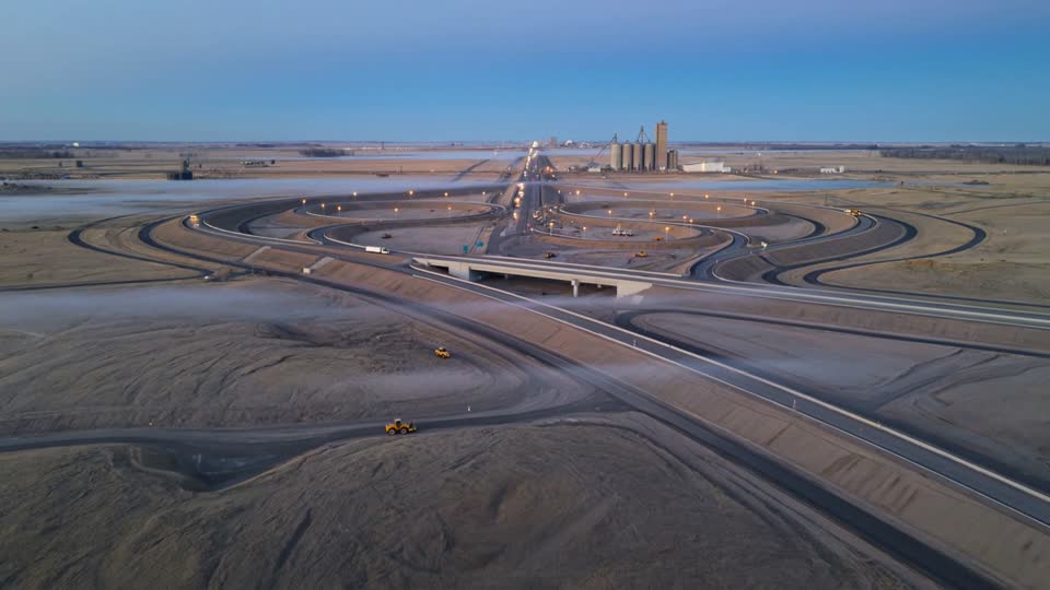 Aerial view at dawn of a highway interchange under construction on the prairie, with curving gravel ramps and grain elevators on the horizon