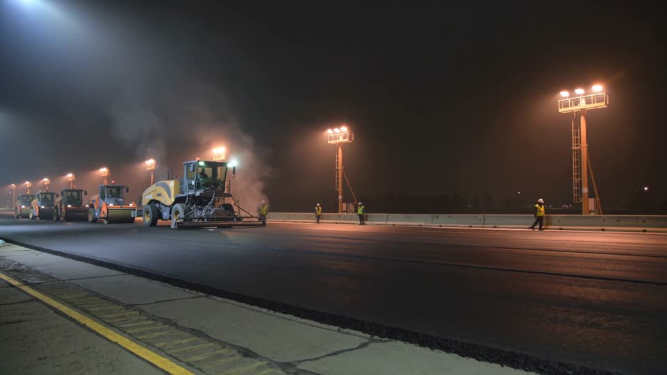 An asphalt paver and rollers laying a steaming mat on a highway at night under light towers