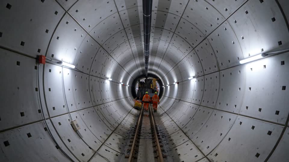Inside a round tunnel lined with precast concrete segments, with a rail track and workers walking toward the boring machine